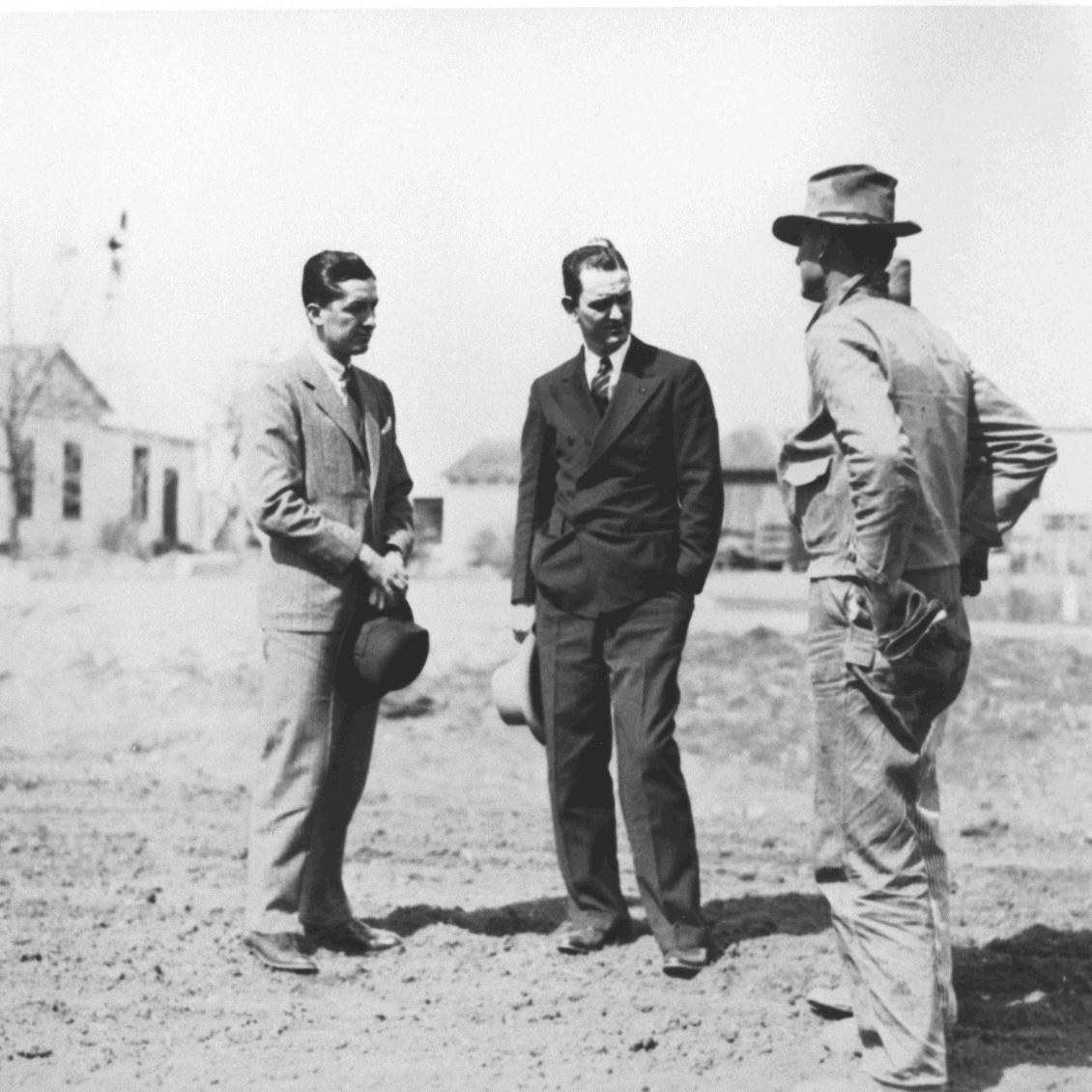 Three men, Lyndon Baines Johnson in center, converse on a dirt roadway.